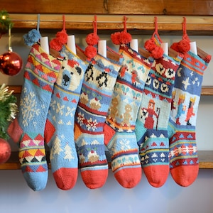 Puede incluir: Una colección de siete calcetines navideños tejidos con varios patrones festivos. Cada calcetín presenta un cuerpo azul claro, una punta roja y un pompón. Los diseños incluyen copos de nieve, pingüinos y formas geométricas, creando un tema navideño acogedor.