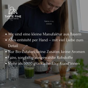 May include: A dark blue t-shirt with the white text TANTE FINE CREW. The image includes text in German about a small Bavarian manufacturer, handmade products, organic ingredients, and over 5000 Etsy customers. A person is whisking green matcha in a bowl.