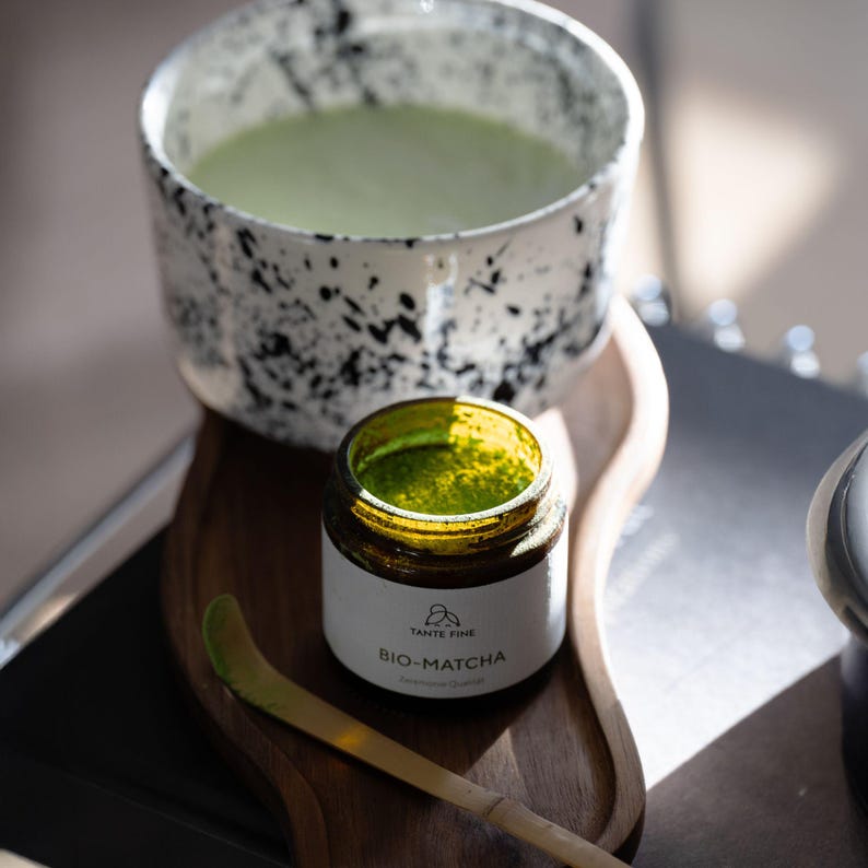 May include: A white ceramic bowl with black speckles containing light green matcha tea, positioned next to a small amber glass jar labeled TANTE FINE BIO-MATCHA and a thin bamboo scoop, all resting on a curved wooden tray surface.