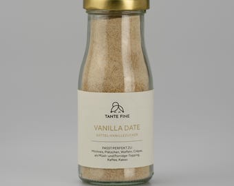 May include: A clear glass bottle with a gold metal lid filled with a light brown granular powder. The white label features the text TANTE FINE, VANILLA DATE, DATTEL-VANILLEZUCKER, and a list of suggested uses including Milchreis, Plätzchen, Waffeln, Crêpes, Müsli, Porridge, Kaffee, and Kakao.
