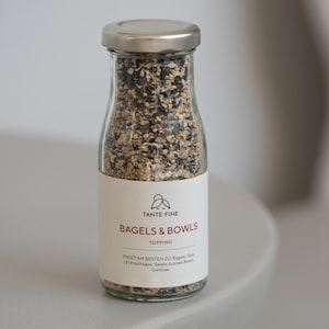 Könnte beinhalten: Eine klare Glasflasche mit silbernem Deckel, gefüllt mit einer Mischung aus Samen und Gewürzen. Das Etikett lautet "TANTE FINE BAGELS & BOWLS TOPPING". Für Bagels, Salate und Gemüse.