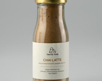 May include: A clear glass bottle with a gold lid filled with a brown powder. The bottle has a white label with the text "TANTE FINE CHAI LATTE" in black. The label also includes German text. The bottle is against a gray background.