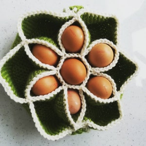 以下が含まれることがあります： 緑と白の編み物でできた花の形をした卵入れ。中に茶色の卵が6個入っています。