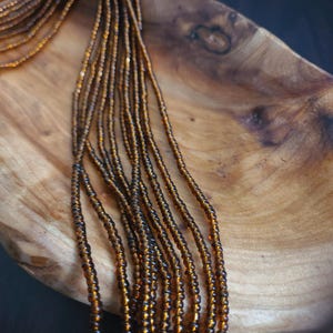 May include: A close-up of a long strand of brown glass beads on a wooden surface. The beads are small and uniform in size, creating a smooth and elegant look.