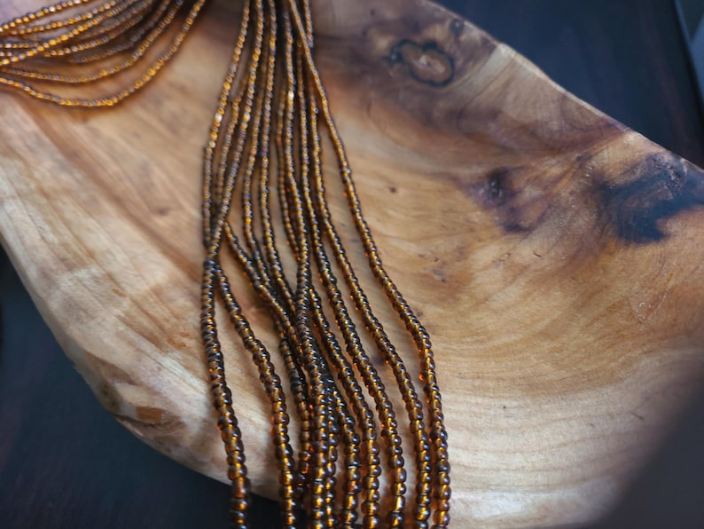 May include: A close-up of a long strand of brown glass beads on a wooden surface. The beads are small and uniform in size, creating a smooth and elegant look.