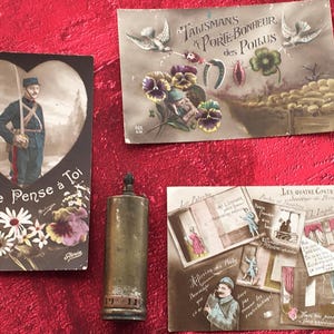 May include: Three vintage postcards with images of a soldier, a horseshoe, and a clover, and a small metal lighter. The postcards have French text, including "Je pense a toi" and "Talisman a Porie Bonheur des Poilus".