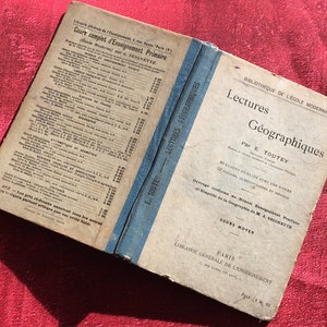 Peut inclure: Un livre vintage intitulé "Lectures Géographiques" par E. Toutey. Le livre fait partie de la série "Bibliothèque de l'Ecole Moderne" et est destiné au niveau "Cours Moyen". La couverture du livre est beige avec du texte bleu et une bande bleue le long de la colonne vertébrale.