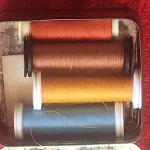 Puede incluir: Una lata de metal con cuatro carretes de hilo. Los carretes son naranja, marrón, amarillo y azul.