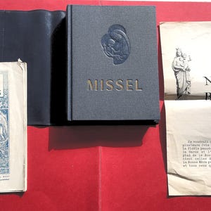 May include: An assortment of religious books and documents on a red surface. A blue-gray book with "MISSEL" in gold lettering is next to a blue book cover. A book titled "LES APPARITIONS DE LOURDES" is also visible, along with a document with the words "Notre Bonne Mère".
