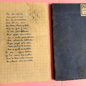 May include: A vintage notebook with a dark blue cover sits next to a handwritten poem on aged, lined paper. The notebook has a label that reads "MABRUG 4CAJ". The poem is in cursive, with visible ink and some smudges.