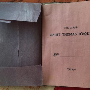 Könnte beinhalten: Ein offenes Buch mit rosa Einband und einem grauen Papierumschlag. Auf dem Einband steht "COURS SAINT THOMAS D'AQUIN" in schwarzer Schrift. Der Umschlag ist über die linke Seite des Buches gefaltet. Das Buch scheint vintage zu sein.