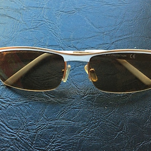 Könnte beinhalten: Sonnenbrille mit weißem Rahmen und braunen Gläsern. Der Rahmen hat goldene Akzente und der Text "UV PROTECTION CE" ist auf dem rechten Bügel aufgedruckt.