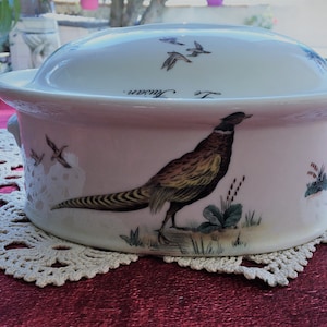 May include: A white and pink ceramic casserole dish with a lid, featuring a pheasant illustration. The dish has two handles and a decorative border. The lid has flying birds and the text "Le Faisan."