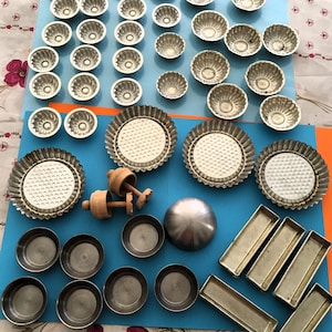 Peut inclure: Une collection de moules et de boîtes de cuisson vintage, comprenant des moules à tartelettes cannelés, des moules à tarte ronds, des petits moules ronds, des moules rectangulaires et une sphère métallique. Les articles sont en métal et en bois, disposés sur une surface bleue.