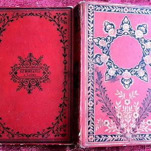 Puede incluir: Libro antiguo abierto con cubiertas rojas y diseños ornamentados en negro y dorado. La cubierta izquierda presenta texto y un borde decorativo. La cubierta derecha tiene un fondo rosa con patrones florales y geométricos.