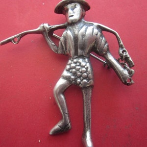 Op de afbeelding: Zilverkleurige figuur van een persoon met een hoed en een stok. De figuur is gedetailleerd met textuur kleding en een ketting.