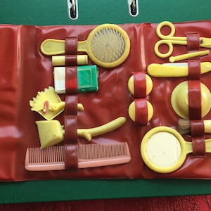May include: A red faux leather travel case containing a variety of yellow plastic toy grooming tools, including a comb, brush, mirror, and razor.