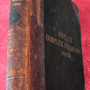 Peut inclure: Un livre relié vintage intitulé Bentley's Complete Phrase Code. Le livre présente une couverture noire foncée avec des lettres dorées et un dos en cuir marron usé et vieilli, montrant des signes évidents d'âge et d'usure du matériau.