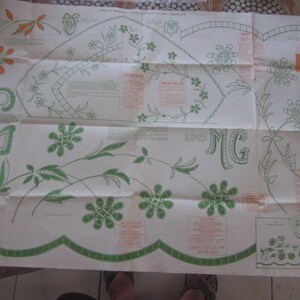 May include: A folded white fabric sheet with green and brown embroidery designs. The sheet has the letters "MG" embroidered in the top right and bottom left corners. The sheet is folded in half and has a scalloped edge.