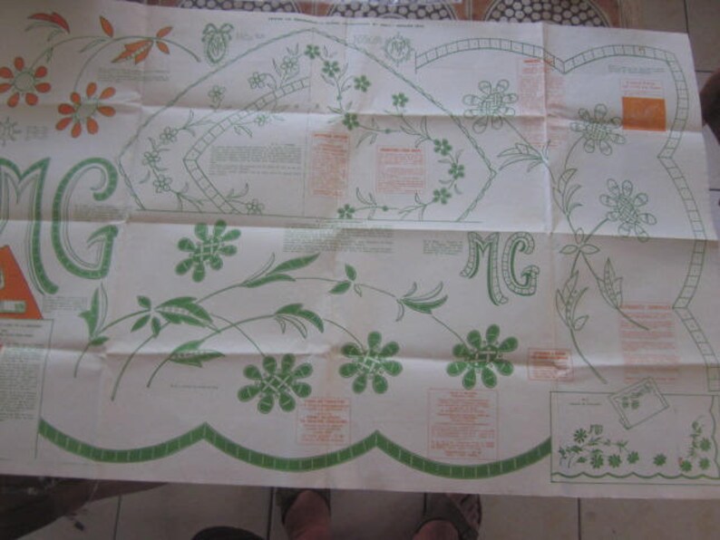 May include: A folded white fabric sheet with green and brown embroidery designs. The sheet has the letters "MG" embroidered in the top right and bottom left corners. The sheet is folded in half and has a scalloped edge.