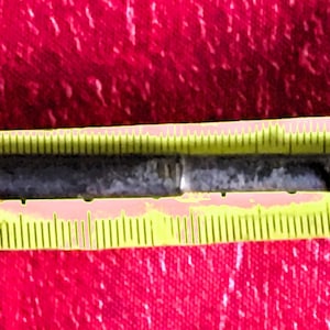 May include: A vintage, dark metal key with a decorative loop attached. The key is approximately 5 inches long, measured against a yellow ruler. The key's teeth are visible, and the metal shows signs of age and wear, suggesting it is an antique.