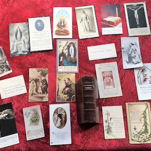 May include: A collection of vintage religious cards and a leather-bound book on a red surface. The cards feature images of saints, religious figures, and scenes from the Bible. The book is titled "Holy Bible" and has a gold-colored spine.