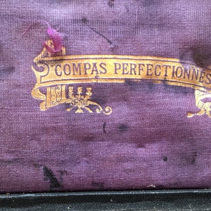 May include: Close-up of a purple velvet box interior with gold lettering that reads "COMPAS PERFECTIONNES". The box has a decorative gold emblem and a purple ribbon. The velvet has dark spots.