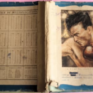 May include: Vintage open book with a blue border. The left page displays a multiplication table. The right page features a painted portrait of a boxer wearing a brown glove. The book's cover is worn and aged.