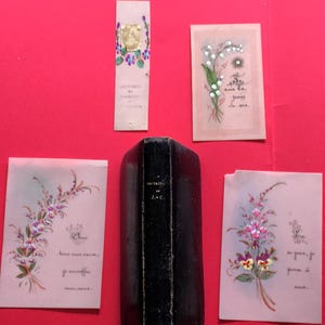 May include: A collection of vintage ephemera, including a small, black leather-bound book and several floral-themed cards. The cards feature delicate watercolor illustrations of flowers and handwritten script. A bookmark with gold accents is also present.