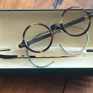 Peut inclure: Lunettes rondes avec une monture à motif écaille de tortue brun foncé et beige et des branches en métal argenté. Les lunettes reposent à l'intérieur d'un étui rigide rectangulaire vert foncé ouvert avec une doublure intérieure beige clair.