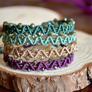 Può includere: Quattro braccialetti con perline impilati su una fetta di legno. I braccialetti sono realizzati con cordoncino verde, turchese, beige e viola e presentano perline di vetro trasparenti.