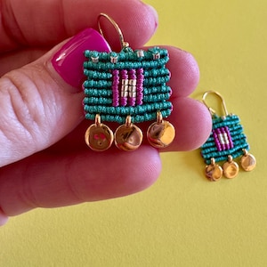 Può includere: Orecchini in macramè fatti a mano con un design intrecciato verde acqua e magenta, accentuati da dettagli in metallo color oro. Ogni orecchino ha una forma quadrata con tre ciondoli circolari pendenti. Su sfondo giallo brillante.