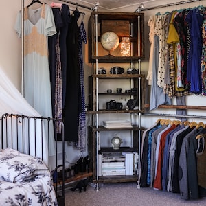 May include: A bedroom with a metal clothing rack and wooden shelving unit. Dresses and shirts hang on the racks. The shelves display cameras, books, and decorative items. A bed with floral bedding is in the foreground.