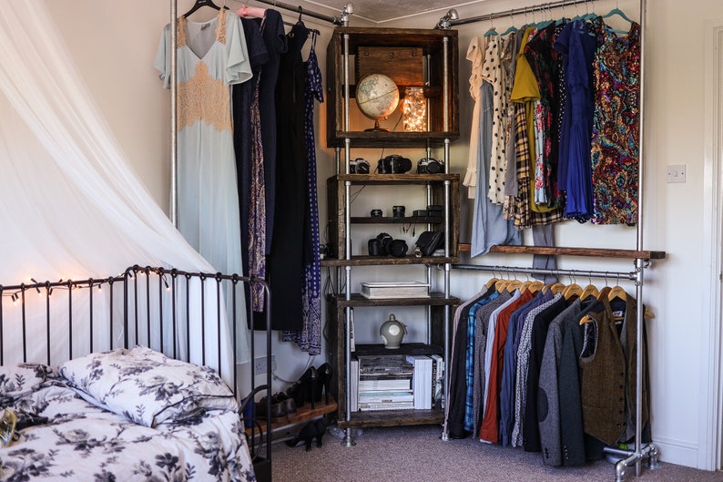May include: A bedroom with a metal clothing rack and wooden shelving unit. Dresses and shirts hang on the racks. The shelves display cameras, books, and decorative items. A bed with floral bedding is in the foreground.