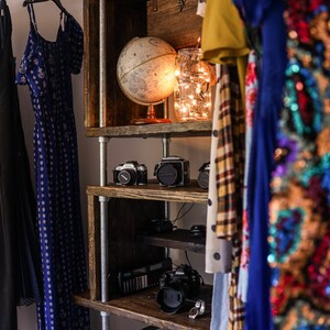 May include: A rustic wooden shelf unit with metal supports, displaying a globe, cameras, and a jar of fairy lights. Clothing hangs on either side, including a blue patterned dress and a sequined garment.