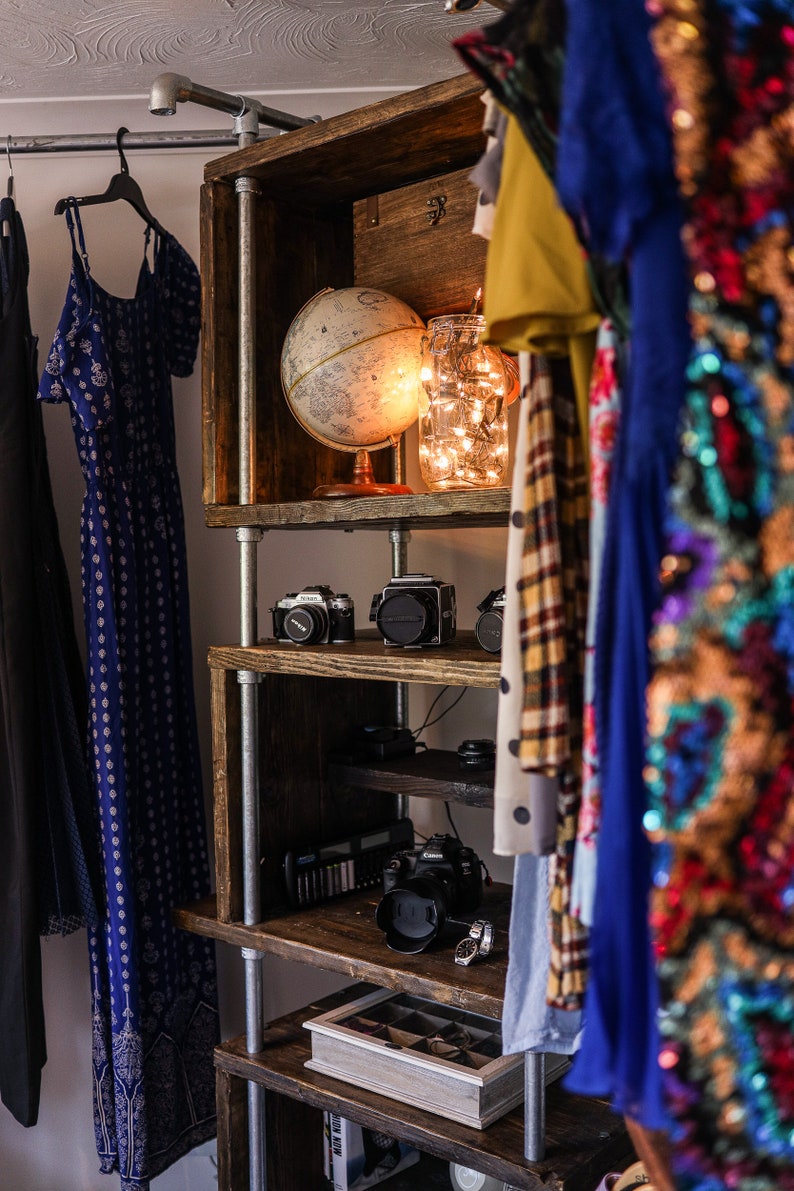 May include: A rustic wooden shelf unit with metal supports, displaying a globe, cameras, and a jar of fairy lights. Clothing hangs on either side, including a blue patterned dress and a sequined garment.