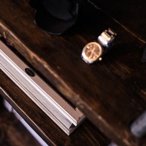 May include: A close-up shot of a dark brown wooden shelf with a level, watch, and other accessories. The level is light gray with a black oval label. The watch has a gold face and a silver band. A black cap and a black leather case are also visible.