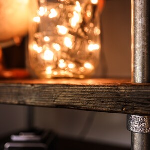 May include: Close-up of a rustic wooden shelf with a metal pipe support. A glass jar filled with glowing fairy lights sits on the shelf, next to a globe. The shelf has a warm, inviting aesthetic, perfect for home decor.