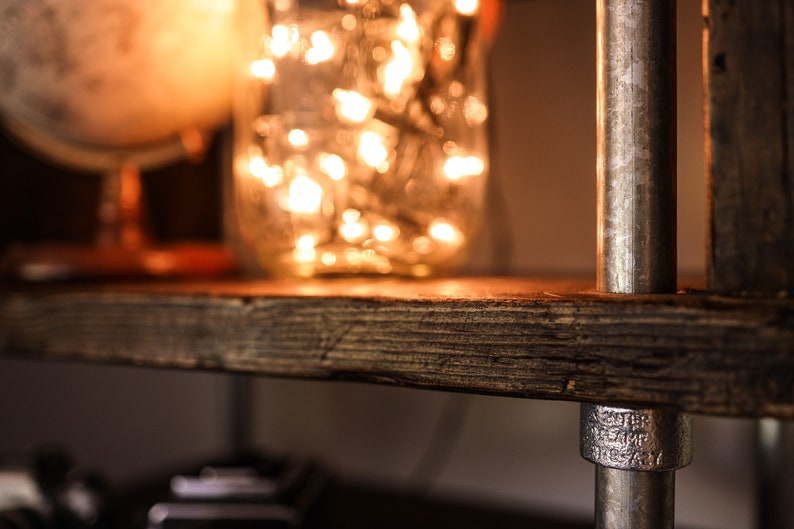 May include: Close-up of a rustic wooden shelf with a metal pipe support. A glass jar filled with glowing fairy lights sits on the shelf, next to a globe. The shelf has a warm, inviting aesthetic, perfect for home decor.