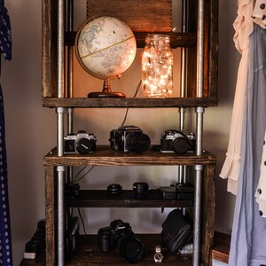 May include: A rustic wooden shelf unit with metal pipe supports. The shelves display a globe, cameras, lenses, and a jar of fairy lights. A small wooden box sits at the top. The unit is in a room with clothing.