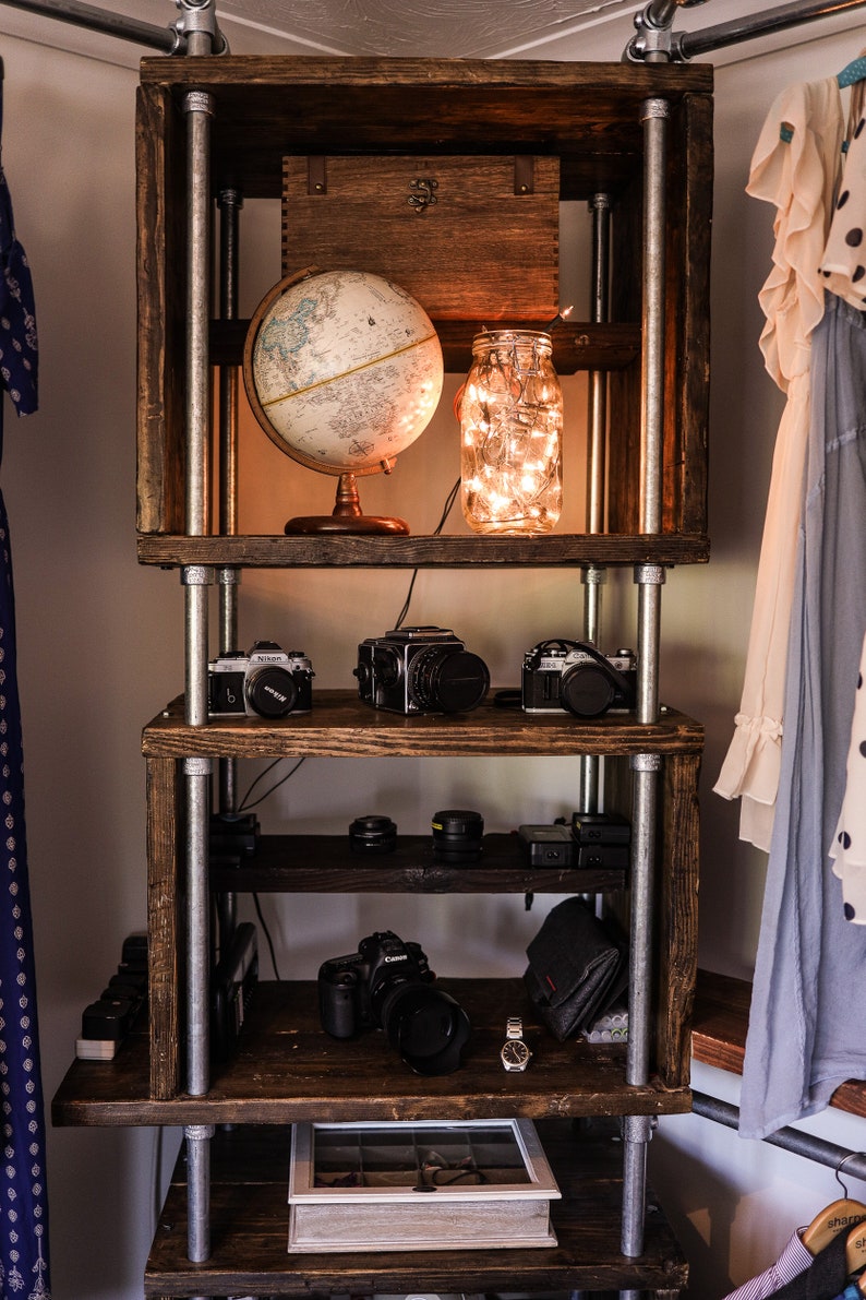 May include: A rustic wooden shelf unit with metal pipe supports. The shelves display a globe, cameras, lenses, and a jar of fairy lights. A small wooden box sits at the top. The unit is in a room with clothing.