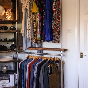 May include: A metal pipe clothing rack with a variety of garments hanging on wooden hangers. The rack holds dresses, shirts, and jackets. A wooden shelf and a wooden shelving unit are visible on the left side of the image.