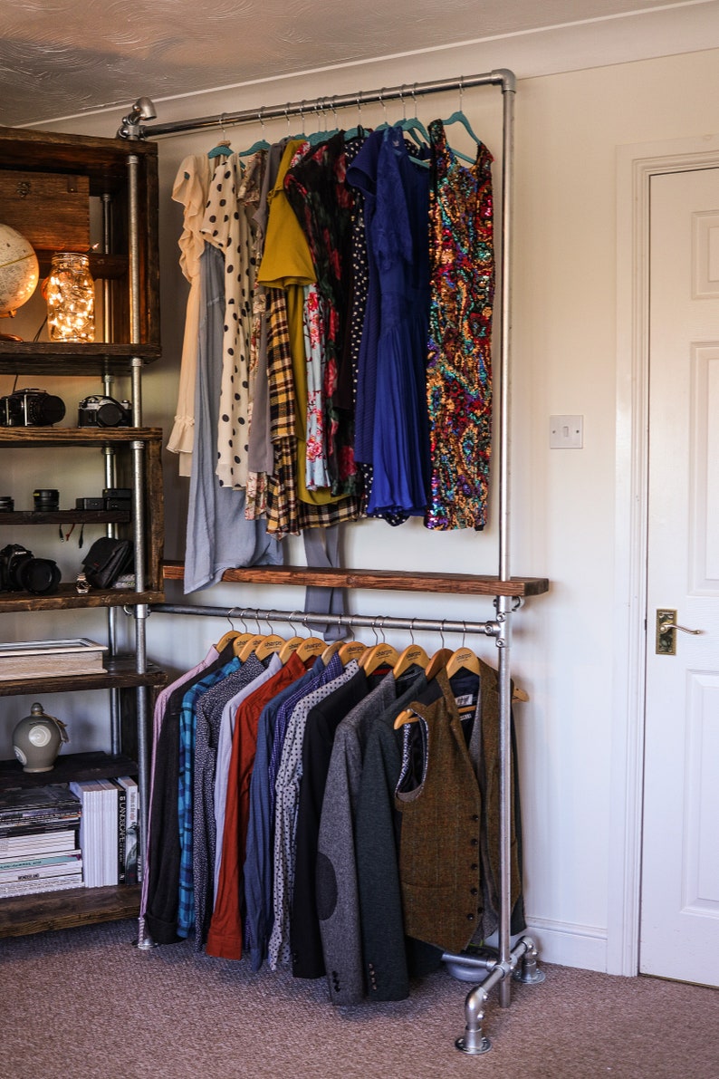 May include: A metal pipe clothing rack with a variety of garments hanging on wooden hangers. The rack holds dresses, shirts, and jackets. A wooden shelf and a wooden shelving unit are visible on the left side of the image.