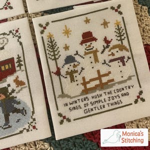 May include: Cross-stitch artwork featuring three snowmen with hats and scarves, a wooden fence, and evergreen trees. The text reads "In Winters Hush the Country Sings, Of Simple Joys and Gentler Things." The artwork is framed with a border of holly.