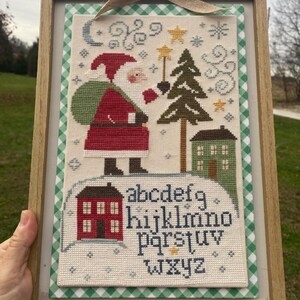 May include: A framed cross-stitch sampler featuring a Santa Claus figure with a green sack, a pine tree, a house, and a star wand. The design includes a red house and the lowercase alphabet letters a through z in dark blue thread on cream fabric.