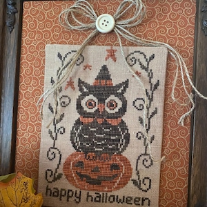 Może przedstawiać: Haft krzyżykowy przedstawiający czarną sowę w kapeluszu czarownicy, siedzącą na pomarańczowej dyni. Poniżej wyhaftowano napis "happy halloween". Dzieło jest oprawione w ciemną drewnianą ramę z dekoracyjną kokardą i guzikiem.