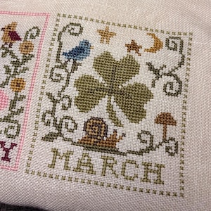 Puede incluir: Bordado de punto de cruz con la palabra "MARZO" en letras beige. El diseño incluye un pájaro azul, un trébol verde de cuatro hojas, un caracol, una seta, estrellas y una luna creciente. Sobre tela beige claro.
