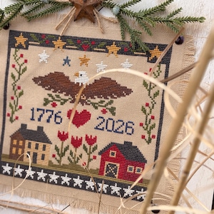 May include: Cross-stitch artwork on burlap fabric, featuring an eagle, stars, and the years 1776 and 2026. The design includes a red heart, houses, and floral elements, framed with a rustic wooden border and decorative greenery.