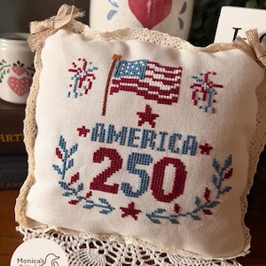 May include: A cream-colored, square pillow with embroidered patriotic design. The design includes an American flag, the word "AMERICA", the number "250", and floral accents in red and blue. The pillow has a lace trim and bow accents.