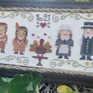Op de afbeelding: Kruissteek sampler met Thanksgiving-figuren: twee inheemse Amerikanen, een kalkoen op een tafel, een pelgrimvrouw en een pelgrimman. De tekst "16 21" staat boven de figuren. De sampler is omlijst met een donkerhouten frame en touwtrim.